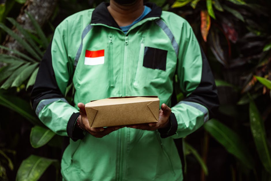 A person dressed in a green and black delivery jacket, featuring a flag detail on the chest, holds a rectangular cardboard box with neatly folded edges in front of lush green foliage. The individual�