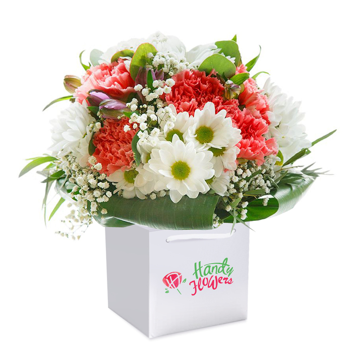 A beautifully orchestrated medley of blooms is showcased upright in a contemporary, square white gift bag, set meticulously against a porcelain white background-a modern touch well-suited to the cosmopolitan charm of Chislehurst. The central ensemble features lush coral and vibrant peach carnations, recognised for their abundant, feathery petals and soft, inviting appearance, clustered in harmonious unity. Scattered among them are pristine white daisies with lime-green hearts, each one radiating cheerful clarity and balancing the warm tones of its carnation companions. Fine-sprayed gypsophila, or baby's breath, laces the bouquet with an airy delicacy, inviting the imagination to envision the tranquil landscapes surrounding Chislehurst Caves. Hinting at life yet to unfurl, a handful of unopened buds tinged in deep violet and pink nestle into the arrangement's left, infusing mystery and anticipation. Enveloping the bouquet is a flourish of glossy, forest-green foliage-broad-leaved and vibrant-creating visual structure and ensuring each bloom claims its own spotlight. This elegant design, enhanced by the clean white gift bag, offers a fresh, ready-to-gift impression ideal for Chislehurst birthdays, anniversaries, or acts of gratitude. The presentation, so lovingly crafted, is a testament to the local florist's expertise and the special care that defines Chislehurst Flowers.