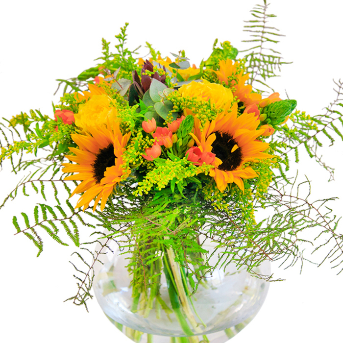 The first impression this bouquet makes is pure, sun-soaked happiness-a tableau of yellow, orange, and green that pays tribute to Chislehurst's open spaces and rolling commons. At the heart are expansive sunflowers, their amber-gold petals splayed wide around cocoa centers, sending out waves of cheerful warmth. Nestled close, lemon-yellow garden roses offer their classic spiral blooms, a nod to cultivated borders and centuries-old gardens lining Chislehurst's High Street. Sprinkled among the blossoms, clusters of deep red hypericum berries glitter, their glossy skins popping against the matte softness of surrounding petals and fronds. Ferns arc gently outward, lending softness and rhythm much like the undulating leafy canopies in Petts Wood. Lighter green filler blooms-perhaps solidago-are woven deftly throughout, giving the bouquet subtle variances in tone and texture. Eucalyptus leaves of cool blue-green introduce an understated, woodland note. All these elements rise and spill artfully from a clear, globed vase, its transparency spotlighting the lush tangle of stems in fresh water-a testament to artisanal care. The bouquet's scent is fresh and uplifting, blending the gentle perfume of cut roses with hints of greenery and countryside air. Thoughtfully composed and deeply local in inspiration, this arrangement is perfect for celebrations, thank-yous, or simply bringing a beam of Chislehurst's natural beauty home.