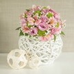 This handcrafted floral centerpiece, set against a gentle, textured beige wall, brings a sense of light and creativity ideal for a modern Chislehurst home or wedding table. The generous bouquet brims with pastel pink and lavender roses, their velvety softness harmonizing with brisk, striped alstroemeria blooms tinged in creamy and fuchsia highlights. Tucked between the flowers, lime green button chrysanthemums capture youthful vibrancy, while clouds of delicate white baby's breath provide an ethereal, floating quality. All are artfully composed in a unique, round white vase of slender, interwoven strips, its open lattice an artistic nod to the craftsmanship of Chislehurst's local artisans. To the left of the bouquet, a pair of pale, rattan-like decorative spheres-one generously sized and grounding, the other smaller and forward-echo the vase's texture, adding balance and a dash of playful elegance to the display. Lit brightly to accentuate each textural detail, the scene reflects the warmth and approachable luxury that define the Chislehurst lifestyle. This arrangement feels at once contemporary and timeless, an expression of joyful celebration, thoughtful gifting, and the heart of community life in Chislehurst.